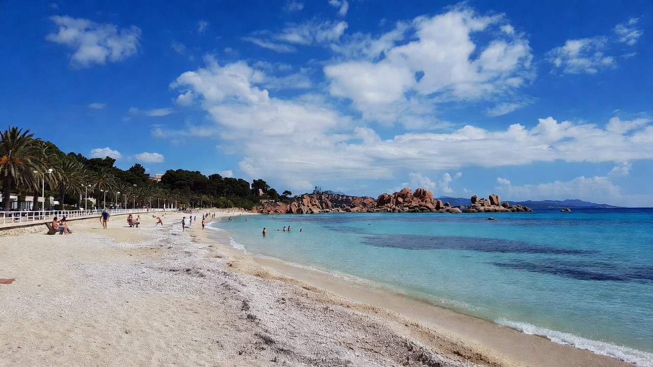 Plage de L’Île-Rousse en Corse avec sable clair, eau turquoise et rochers rouges, située près du centre-ville, montrant l’accès facile, les zones de parking à proximité et une ambiance familiale.